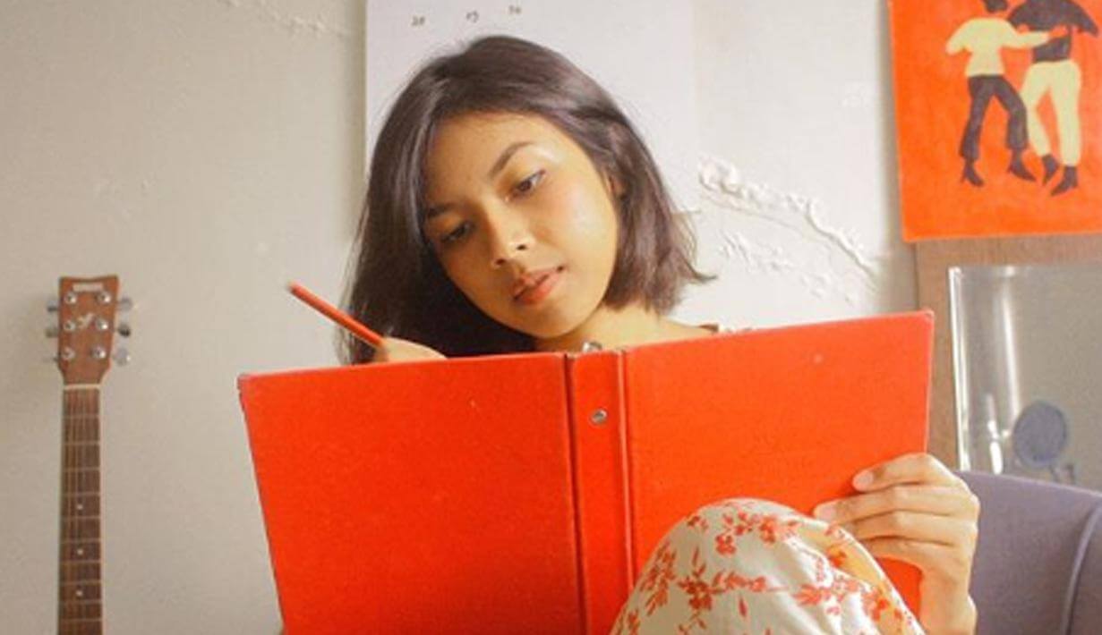 Vira Talisa sedang duduk di sofa sambil menulis di buku berwarna orange